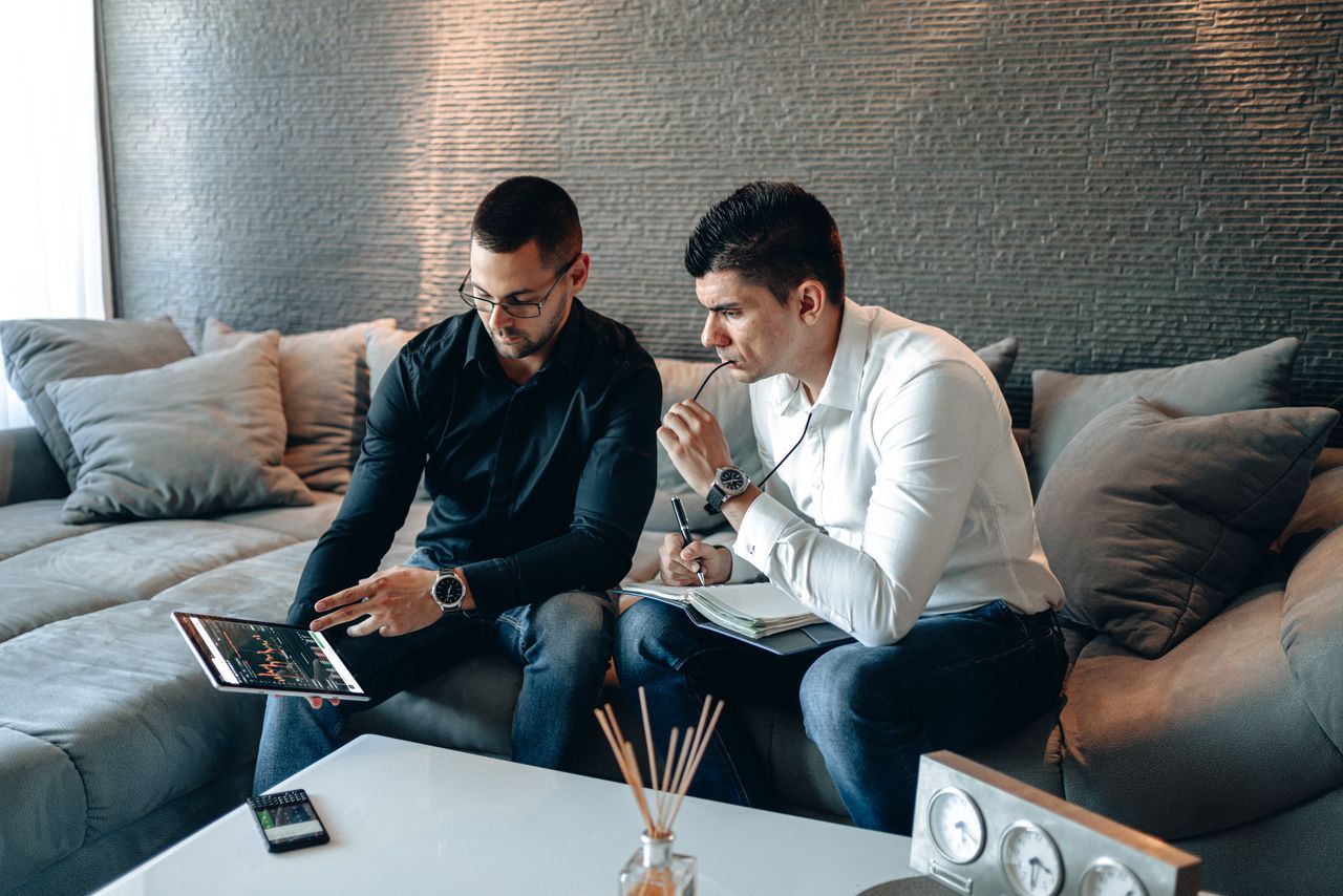 hero-img-02 Two business professionals on a sofa analyzing financial graphs on a digital tablet indoors.